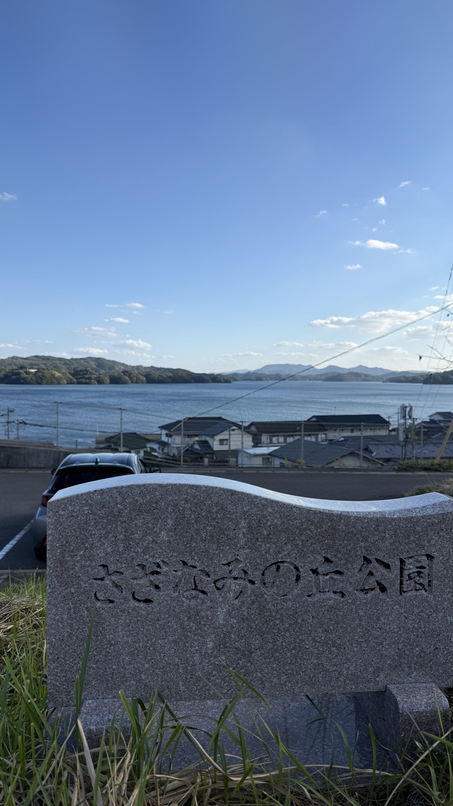 「さざなみの丘公園」ときざまれた石碑。そのむこうに家なみと形上湾、対岸の山がひろがる