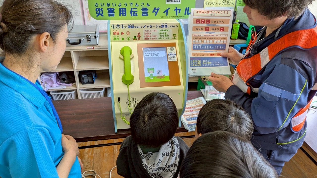 災害用伝言ダイヤルの体験。電話の模型の前で、子どもたちが係の人の説明を聞いている