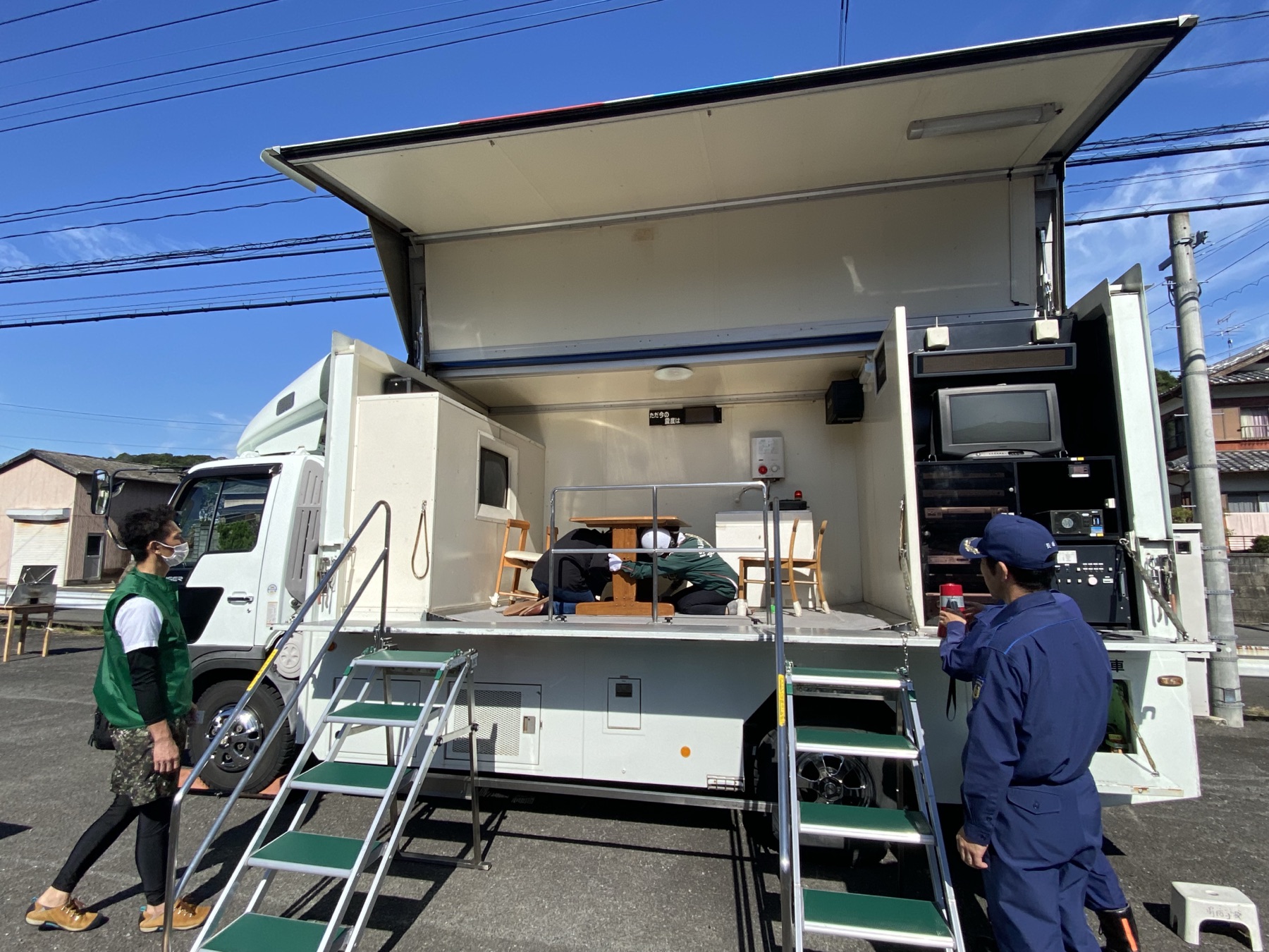 地震体験車のまわりに集まる人たち。荷台では机の下に身をかがめて揺れにそなえている